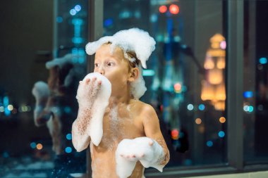Akşam şehir manzaralı bir penceresi arka plan üzerinde uyumaya gitmeden önce akşam banyo küvete oturan mutlu küçük oğlu. Bir banyoda pencere yakınındaki köpük dolu banyo bebek portresi.