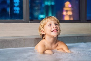 Akşam şehir manzaralı bir penceresi arka plan üzerinde uyumaya gitmeden önce akşam banyo küvete oturan mutlu küçük oğlu. Bir banyoda pencere yakınındaki köpük dolu banyo bebek portresi.