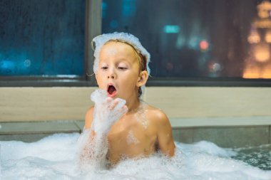 Akşam şehir manzaralı bir penceresi arka plan üzerinde uyumaya gitmeden önce akşam banyo küvete oturan mutlu küçük oğlu. Bir banyoda pencere yakınındaki köpük dolu banyo bebek portresi.