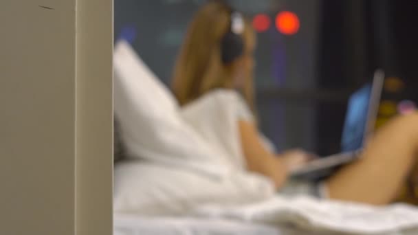 Mujer joven con auriculares inalámbricos trabajando con un portátil sentado en su cama con siluetas de rascacielos en un backgound — Vídeos de Stock