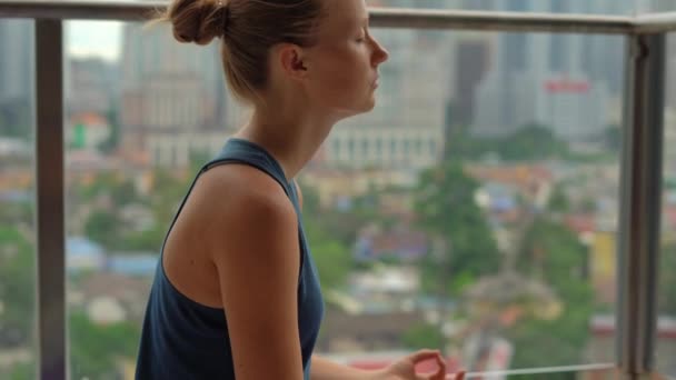 Junge Frau macht Yoga-Übungen auf ihrem Balkon in einem mehrstöckigen Gebäude mit Blick auf eine Innenstadt mit Wolkenkratzern. Geistiges Gleichgewicht in einem Großstadtkonzept — Stockvideo