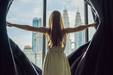 Genç kadın pencere perdeleri açar ve büyük şehirde gökdelenler bakar.