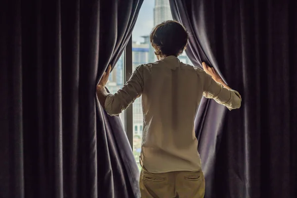 Young man opens the window curtains and looks at the skyscrapers in the big city.