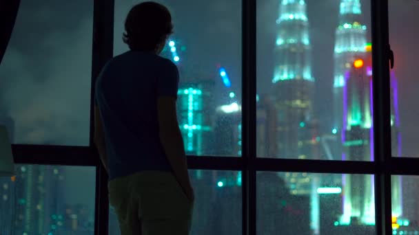 Силуэт человека, стоящего у панорамного окна ночью и смотрящего на небоскребы в центре города — стоковое видео
