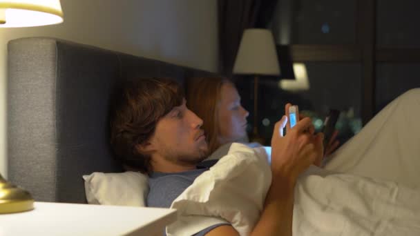 Чоловік і жінка лежать в ліжку кожен зі смартфоном і планшетним ПК. проблеми у шлюбі та міжстатевих відносинах та концепція залежності мобільних пристроїв — стокове відео