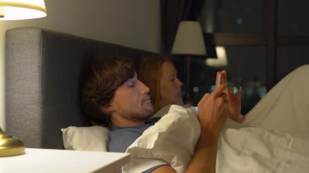 Homme et femme couchés dans le lit chacun avec smartphone et tablette pc. L'homme éteint la lampe et s'endort alors que la femme ne l'éteint pas. problèmes de mariage et de relations intersexuelles problèmes et mobiles — Video