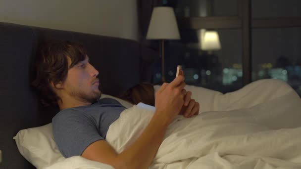 Мужчина и женщина лежат в постели, мужчина со смартфоном, пока женщина спит. проблемы в браке и интерсексуальные проблемы отношений и мобильные устройства наркомании концепции — стоковое видео
