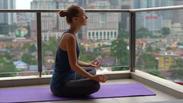 Mujer joven haciendo ejercicios de yoga en su balcón en un edificio de varios pisos con vistas a un centro de la ciudad con rascacielos. Equilibrio mental en una gran ciudad — Vídeo de stock