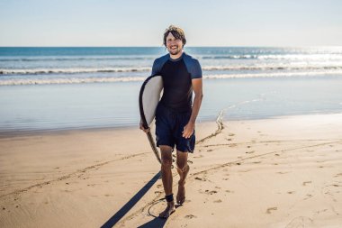 Yakışıklı sportif genç sörfçü tropikal bir kumsalı üzerinde onun ıslak elbiseli kolunun altında onun surfboard ile poz.