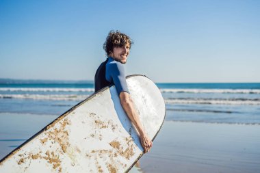 Yakışıklı sportif genç sörfçü tropikal bir kumsalı üzerinde onun ıslak elbiseli kolunun altında onun surfboard ile poz.