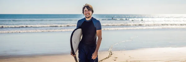 Handsome sporty young surfer posing with his surfboard under his arm in his wet suit on a sandy tropical beach.