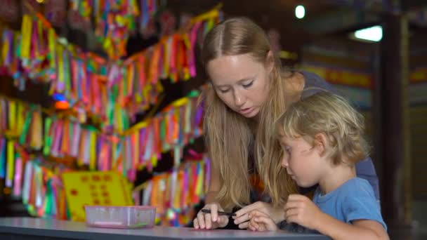 Eine junge Frau und ihr kleiner Sohn besuchen den buddhistischen Tempel kek lok si und schreiben Wünsche auf ein farbiges Textil — Stockvideo