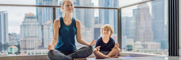 Mom and son are practicing yoga on the balcony in the background of a big city. Sports mom with kid doing morning work-out at home. Mum and child do the exercises together, healthy family lifestyle concept. BANNER, long format