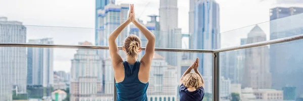 Mom and son are practicing yoga on the balcony in the background of a big city. Sports mom with kid doing morning work-out at home. Mum and child do the exercises together, healthy family lifestyle concept. BANNER, long format