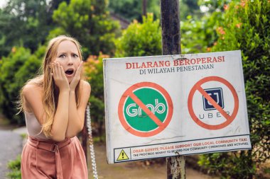 BALI, INDONESIA - May 21, 2018: Young woman looking at protest sign on wall in Indonesian objecting to Uber and Grab taxi drivers reading 'Uber and Grab Taxis No Entry'.