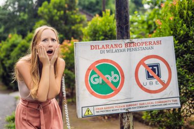 BALI, INDONESIA - 21ST MAY, 2018: Young woman looks at protest sign on a wall in Indonesian objecting to Uber and Grab taxi drivers reads 'Uber and Grab Taxis No Entry' in Ubud, Bali, Indonesia. clipart