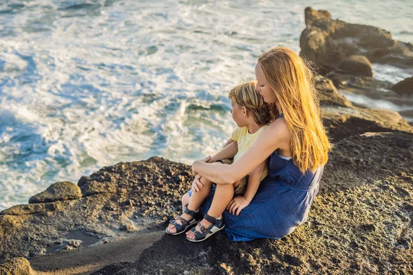 Mom and son are sitting on a rock and looking at the sea. Portrait travel tourists - mom with kids. Positive human emotions, active lifestyles. Happy young family on sea beach.