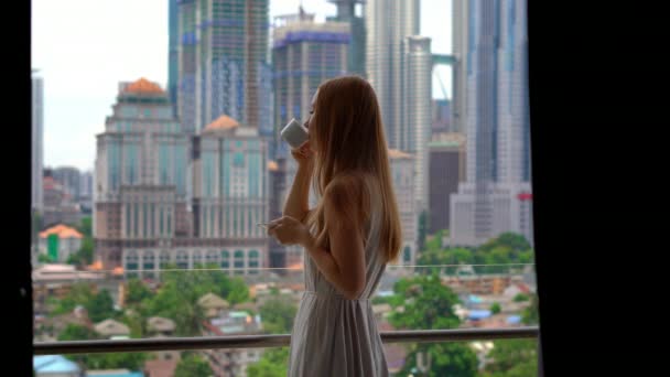 Jeune femme sur un balcon de son appartement avec vue sur un centre-ville plein de gratte-ciel boit du café le matin . — Video