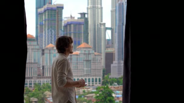 Jongeman komt een balkon van zijn appartement met uitzicht op een centrum van de stad vol skyscrappers en drinkt zijn koffie ochtend — Stockvideo