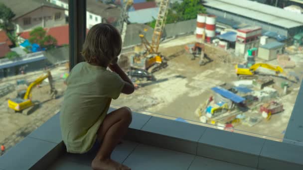 Ein kleiner Junge sitzt auf einem Balkon mit Glaswänden und beobachtet den Bau eines mehrstöckigen Gebäudes — Stockvideo