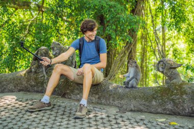 Genç adam komik makak maymunu Bali ormanda ile selfie üstündə selfie stick kullanarak.