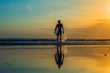 deniz kıyısı boyunca surfboard ile günbatımında yürüyen genç adam