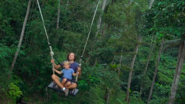 Медленный снимок молодого человека и его сына, качающихся на больших качелях над скалой в тропиках — стоковое видео