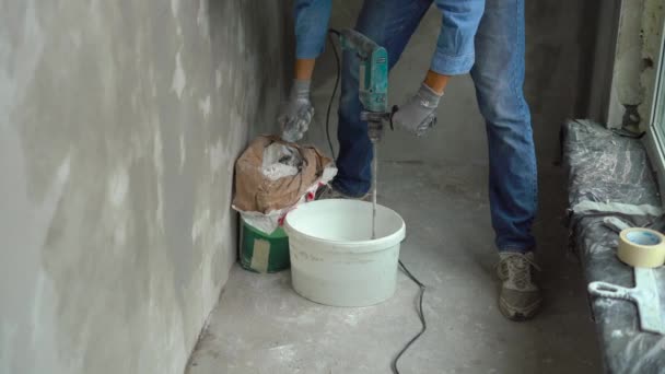 A young male painter kneads putty with water in a bucket using a hand-held mixer for building mixes — Stock Video