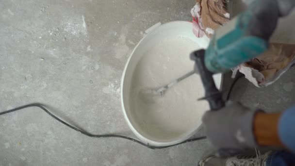 A young male painter kneads putty with water in a bucket using a hand-held mixer for building mixes — Stock Video