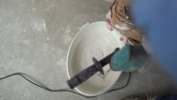 Un joven pintor amasa masilla con agua en un cubo usando un mezclador de mano para construir mezclas — Vídeo de stock