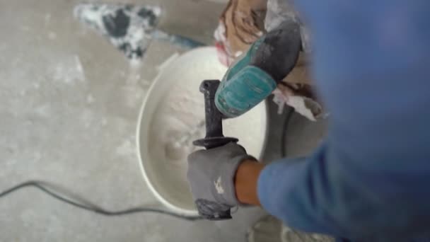 Un joven pintor amasa masilla con agua en un cubo usando un mezclador de mano para construir mezclas — Vídeos de Stock
