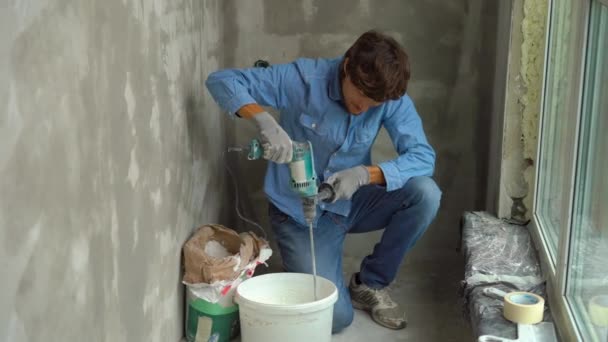 A young male painter kneads putty with water in a bucket using a hand-held mixer for building mixes — Stock Video