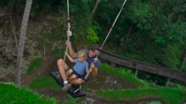 Genç bir adam ve oğlu tropikal bir uçuruma büyük salıncakta sallanan Slowmotion atış