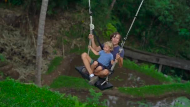 Slowmotion shot of a young man and his son swinging on a large swing over a cliff in the tropics — Stock Video