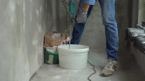 A young male painter kneads putty with water in a bucket using a hand-held mixer for building mixes — Stock Video