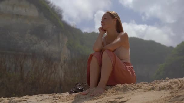 バリ島メラスティ ビーチで背景の岩と砂の上に座っていた若い女性 — ストック動画