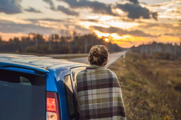 Una mujer en una manta a cuadros está al lado del coche al lado de la carretera en el fondo del amanecer. Concepto de viaje por carretera — Foto de Stock