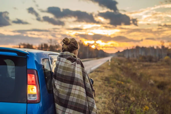 Una donna in un plaid si trova accanto alla macchina sul ciglio della strada sullo sfondo dell'alba. Concetto di viaggio — Foto Stock