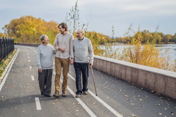 Een bejaarde echtpaar wandelingen in het park met een mannelijke assistent of volwassen kleinzoon. Zorg voor ouderen, vrijwilligerswerk — Stockfoto