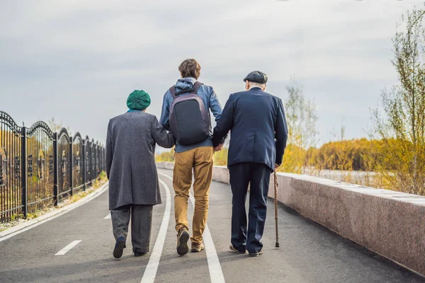 Vanhempi pari kävelee puistossa miespuolisen avustajan tai aikuisen pojanpojan kanssa. Vanhustenhoito, vapaaehtoistyö — kuvapankkivalokuva