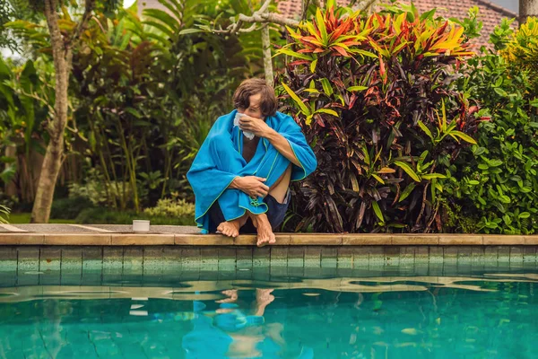 Viajante doente. O homem pegou um resfriado nas férias, senta-se triste na piscina bebendo chá e sopra o nariz em um guardanapo. Seu filho está saudável e nadando na piscina. Conceito de seguro de viagem — Fotografia de Stock