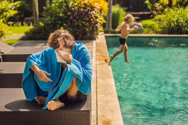 Kranker Mann. Der Mann hat sich im Urlaub erkältet, sitzt traurig am Pool und trinkt Tee und pustet seine Nase in eine Serviette. Sein Sohn ist gesund und schwimmt im Pool. Reiseversicherungskonzept — Stockfoto