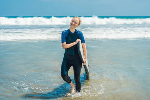 Smiling blonde woman in swimsuit with surf for beginners ready to surf in ocean water.