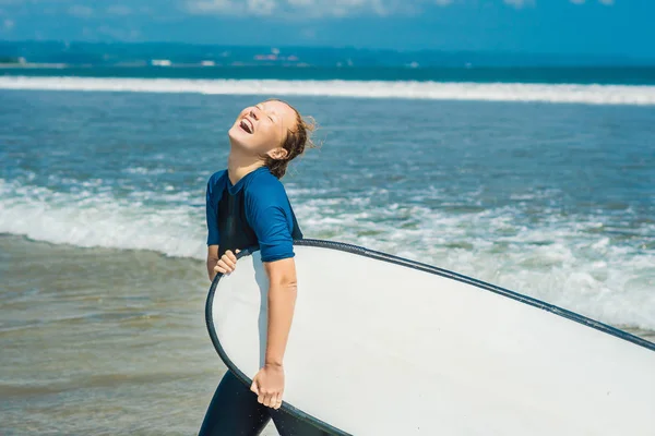 Laughing blonde woman in swimsuit with surf for beginners ready to surf in ocean water.