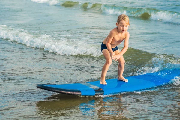 Little boy learning to surf on blue board at tropical beach.