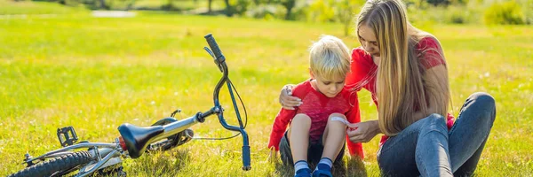 El niño se cayó de la bicicleta, su madre pega un yeso en su rodilla BANNER, FORMATO LARGO — Foto de Stock