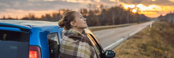 Une femme en plaid se tient près de la voiture sur le bord de la route en arrière-plan de l'aube. Concept de road trip BANNER, FORMAT LONG — Photo