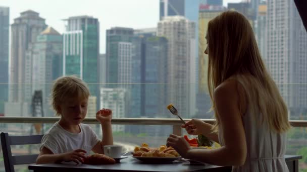 Jonge vrouw gouvernante ontbijten met een jongen op een balkon met uitzicht op de wolkenkrabbers van het centrum van de stad — Stockvideo