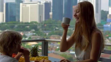Genç kadın mürebbiye şehir merkezini gökdelenler bakan bir balkon bir çocukla kahvaltı var