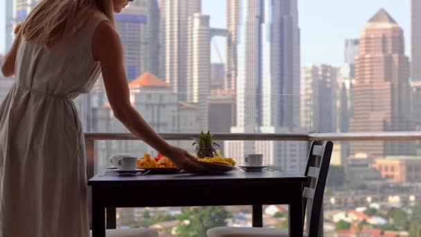 Jovem governanta preparando café da manhã em uma varanda com vista para os arranha-céus do centro da cidade — Vídeo de Stock